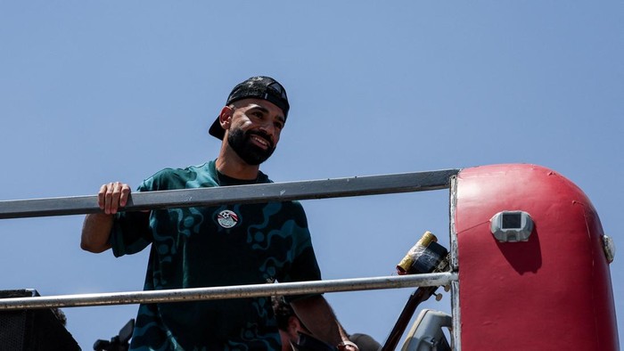 Smiling man with a backward cap leaning on a metal railing of a parade float, blue sky behind and a red inflatable barrier nearby.