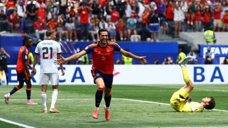 Soccer scene: a player in a red Spain jersey runs with arms spread, celebrating near a goalkeeper lying on the turf in a yellow kit as fans cheer.