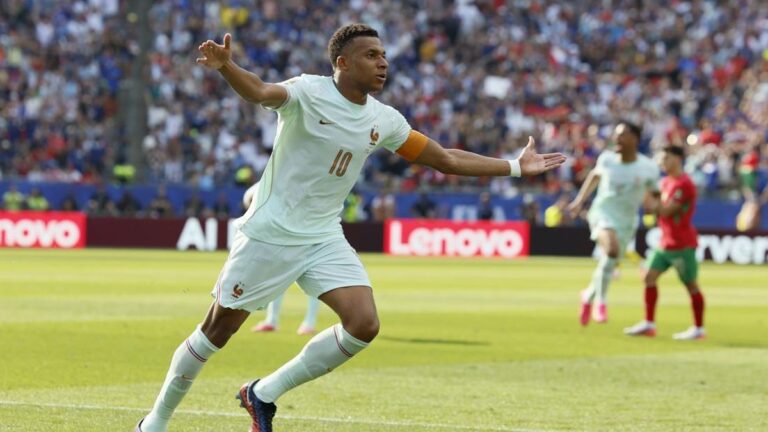French footballer with number 10 shirt celebrates a goal, arms outstretched on a sunny pitch with fans in the stands behind him.