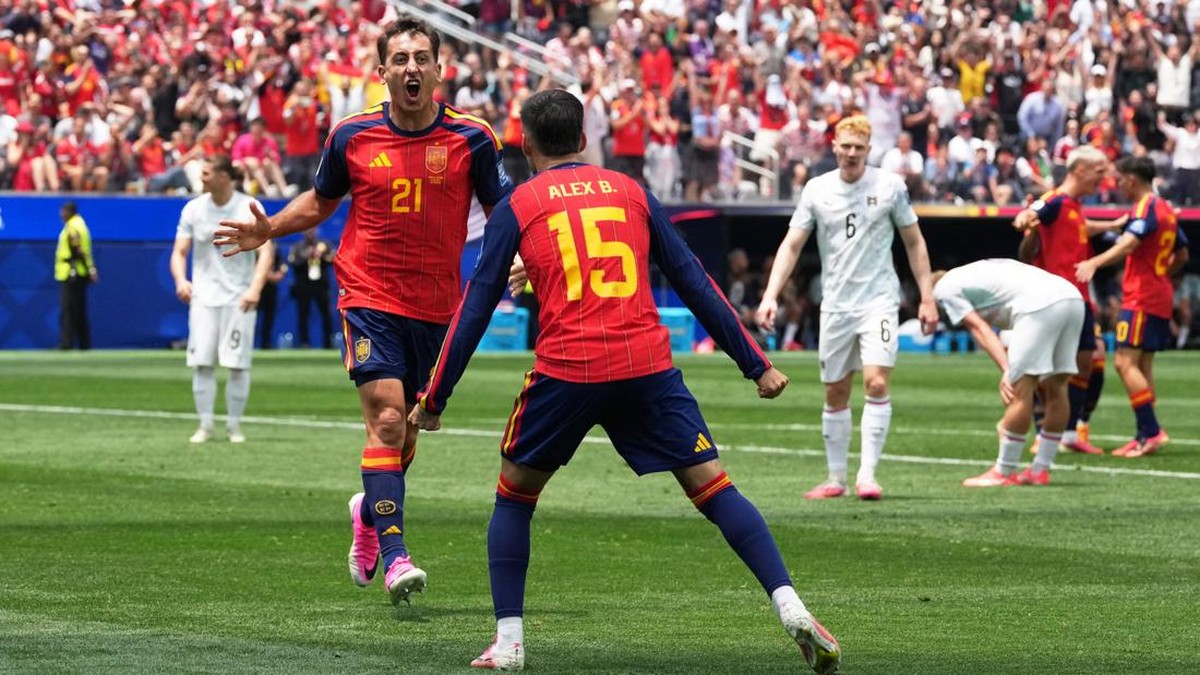 Spain players in red jerseys celebrate a goal on a sunny, crowded stadium field; number 21 is cheering while teammate number 15 (Alex B.) approaches with arms outstretched, white-shirted opponents in the background.