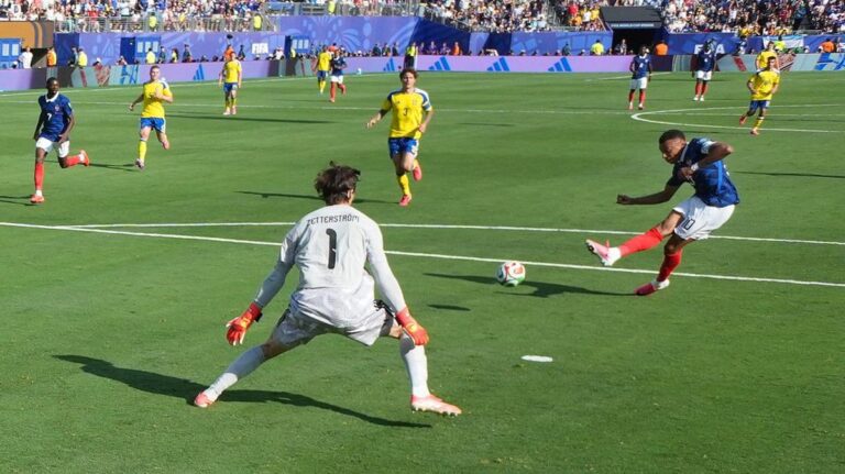 Soccer scene: a blue-team player kicks the ball toward goal while the white-clad goalkeeper (#1) готовs to block, with yellow-team players and a crowded stadium in the background.