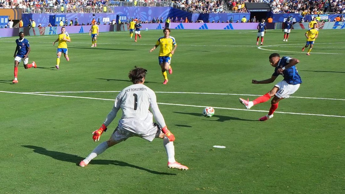 Soccer scene: a blue-team player kicks the ball toward goal while the white-clad goalkeeper (#1) готовs to block, with yellow-team players and a crowded stadium in the background.