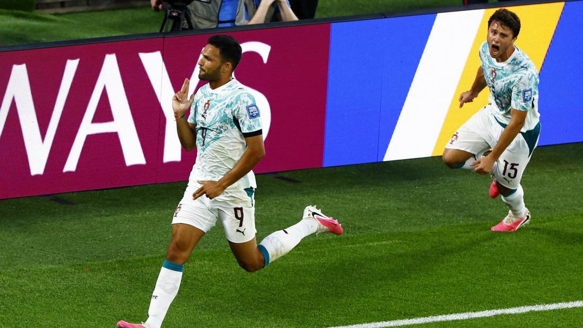 Two soccer players in light-blue jerseys celebrate a goal, sprinting on a green field near advertising boards.