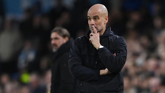 Bald man in a dark jacket on a sideline, finger to lips in a 'quiet' gesture during a football match