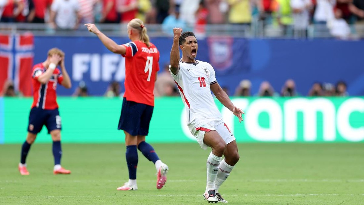 England footballer in white jersey #10 celebrates a goal with a raised fist on the pitch, red-clad teammates nearby and a cheering crowd behind.