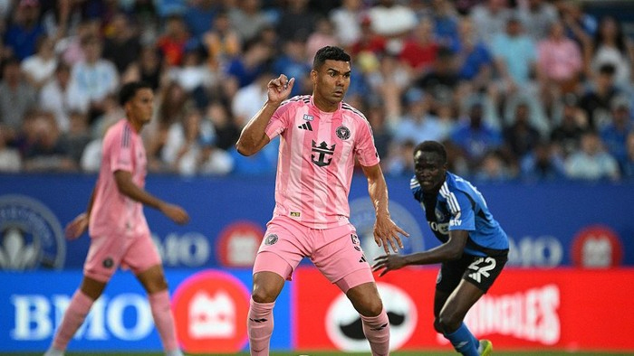 Soccer player in a pink kit dribbles the ball during a match, with a blue-clad opponent nearby and a crowded stands behind him.
