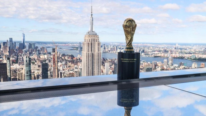 FIFA World Cup trophy on a pedestal, reflected in a glass surface, with the Manhattan skyline and Empire State Building in the background.