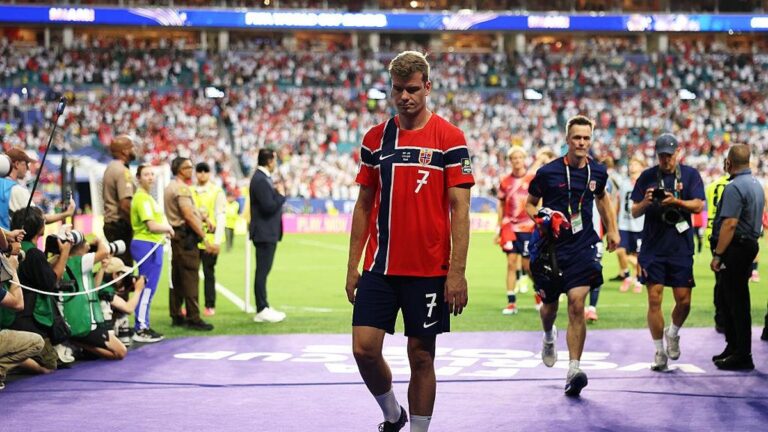 Soccer player wearing a red jersey with number 7 walks off the field as photographers and staff line the sideline and a full stadium watches.