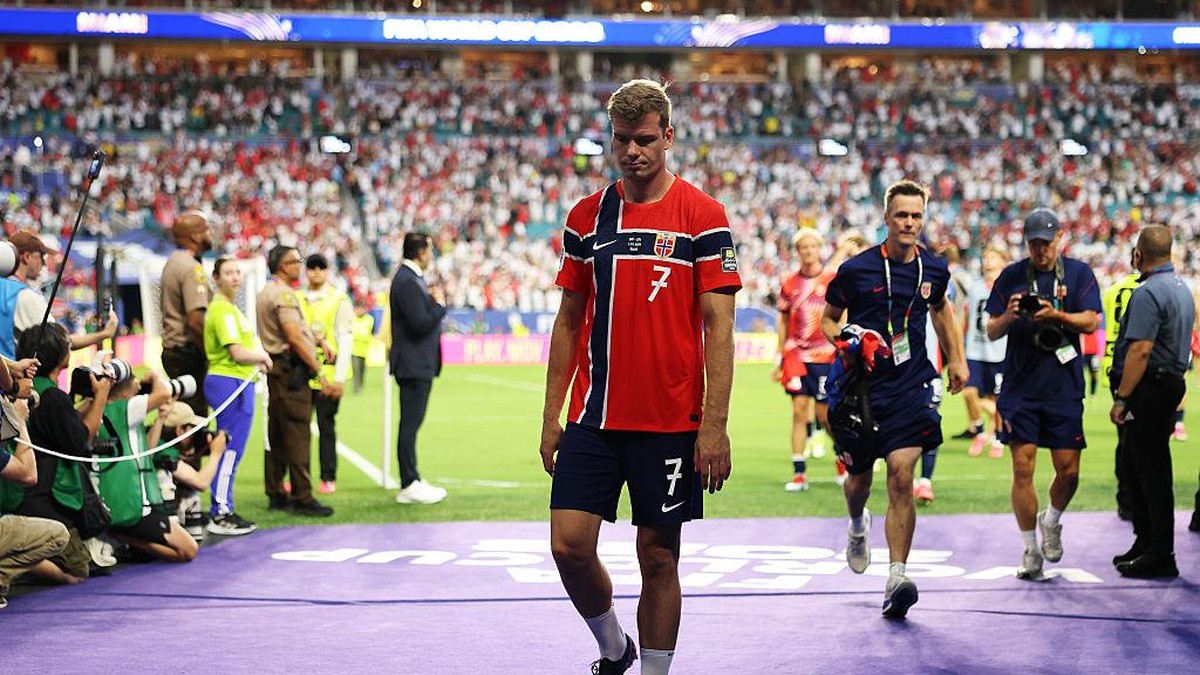Soccer player wearing a red jersey with number 7 walks off the field as photographers and staff line the sideline and a full stadium watches.