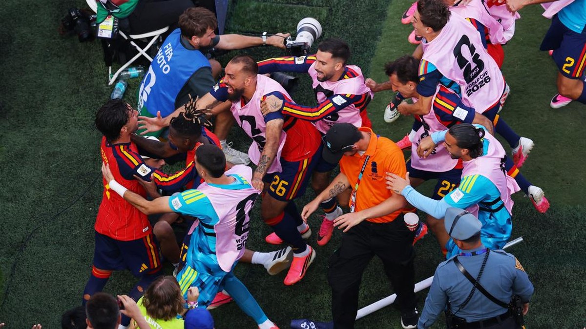 Soccer players in pink bibs celebrate together on a green field, surrounded by photographers and staff in a post‑match huddle.