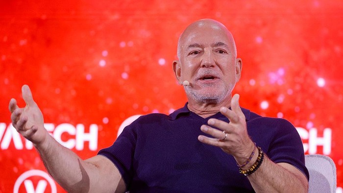 Speaker with a headset microphone gesturing with hands on a red stage backdrop at a conference.