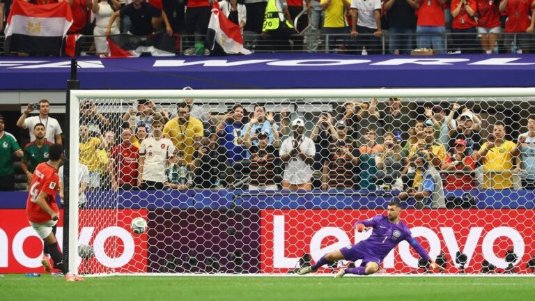 Soccer goalkeeper in purple dives to block a low shot near the goal as a red-clad attacker approaches; crowd cheers behind the net.