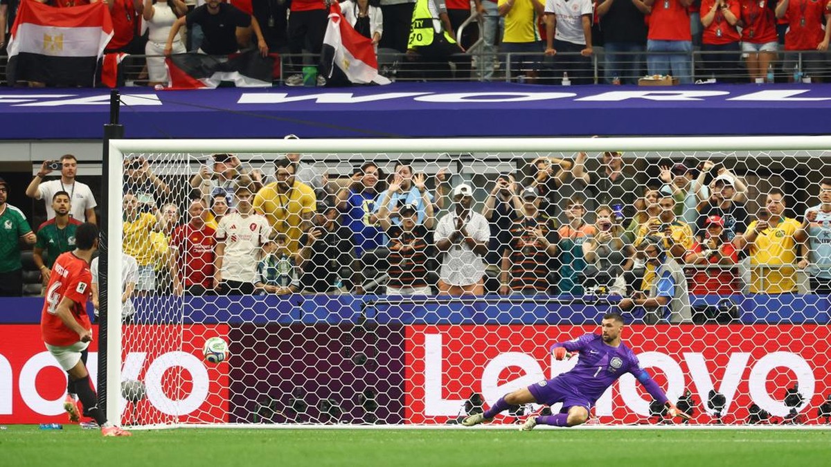 Soccer goalkeeper in purple dives to block a low shot near the goal as a red-clad attacker approaches; crowd cheers behind the net.
