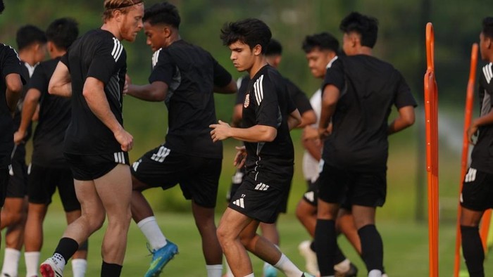Group of soccer players in black kits sprint during a training drill on a green field with orange training poles nearby.