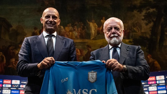 Two men in suits smile while presenting a blue soccer jersey on a stage with a banner in the background.