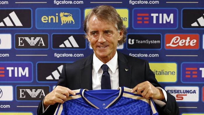 Man in a suit holding up a blue soccer jersey at a backdrop covered with sponsor logos during a press conference.