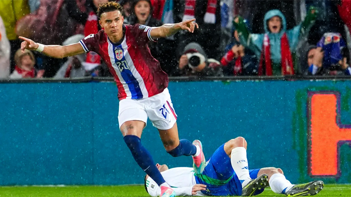 Soccer player in a red-and-blue striped jersey leaps over a sliding opponent on a wet, rain-soaked pitch with cheering fans in the stands.