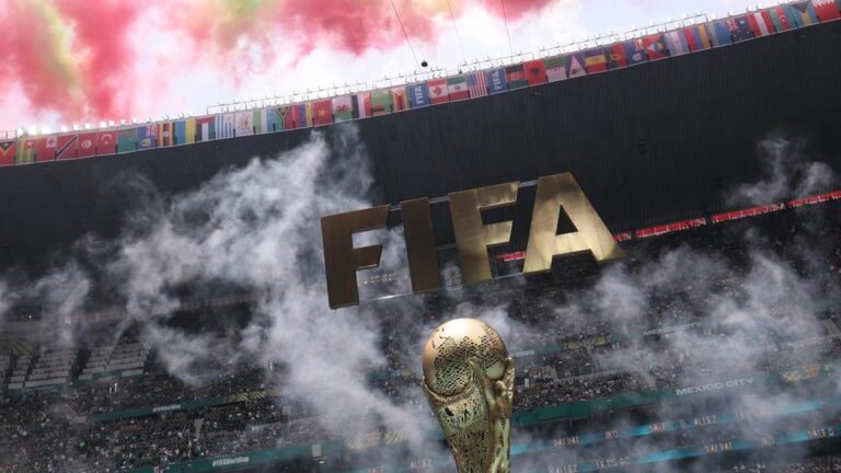 FIFA World Cup trophy on the field with oversized FIFA letters above and colorful flags, red smoke in the stands.