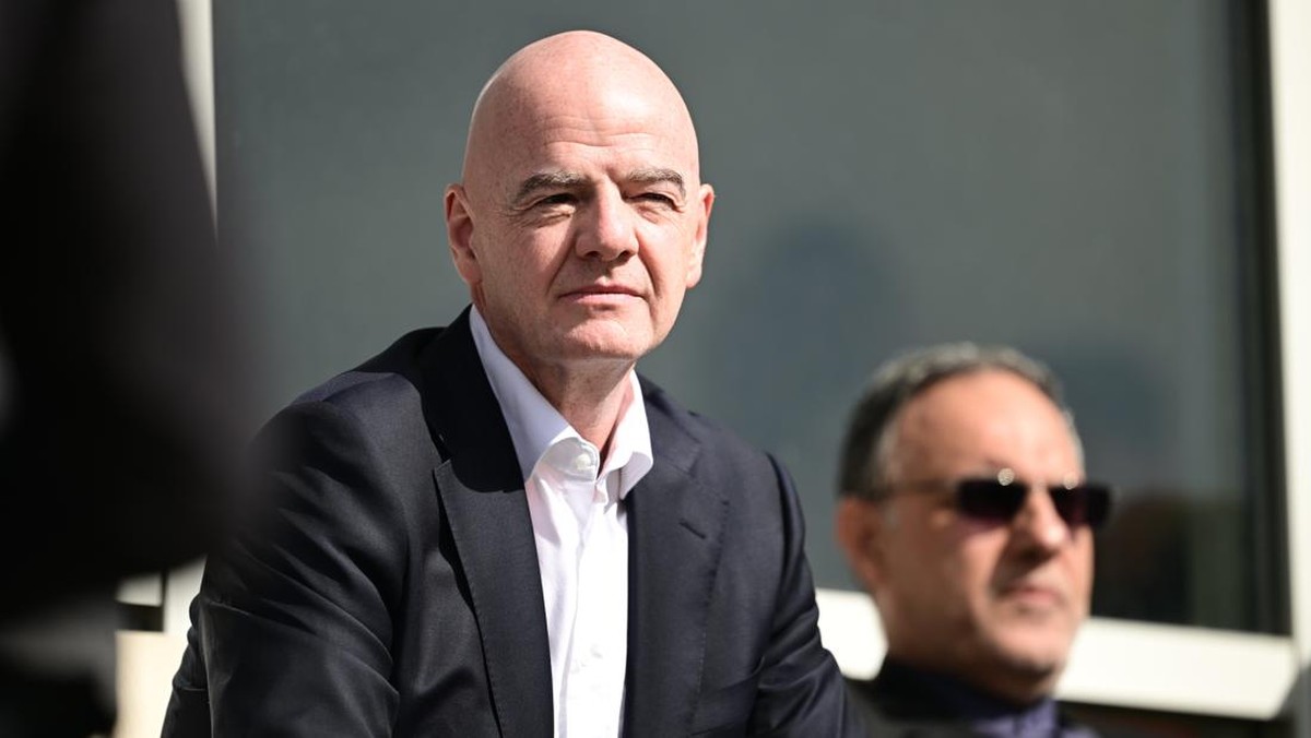 Bald man in a dark suit and white shirt sits outdoors, with a man in sunglasses blurred in the background at a business event.