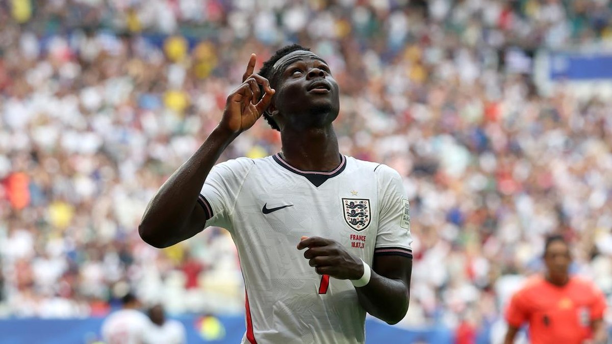 England footballer in a white kit points upward in celebration with a packed stadium behind him, after a goal.