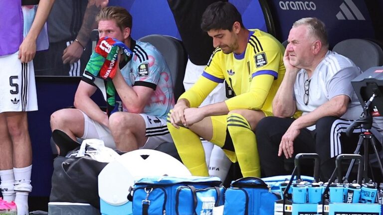 Soccer players resting on a bench: one keeper wipes his face with a towel, another in yellow kit sits with hands together, an older man watches nearby as gear and water bottles sit in front.
