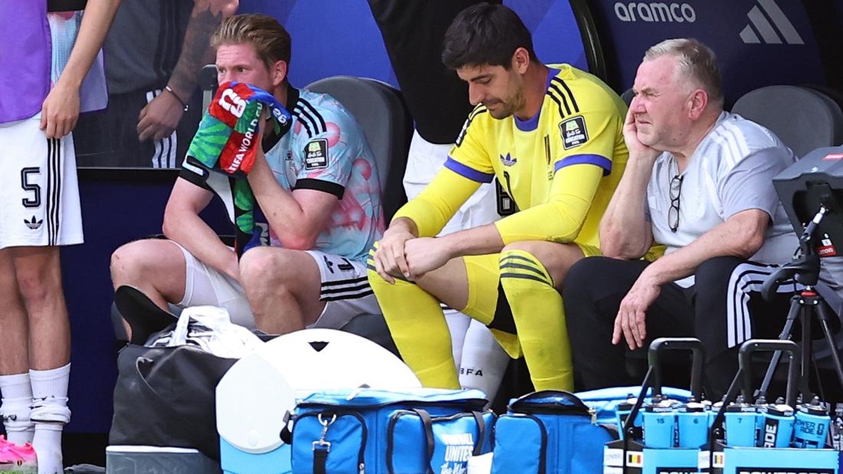 Soccer players resting on a bench: one keeper wipes his face with a towel, another in yellow kit sits with hands together, an older man watches nearby as gear and water bottles sit in front.