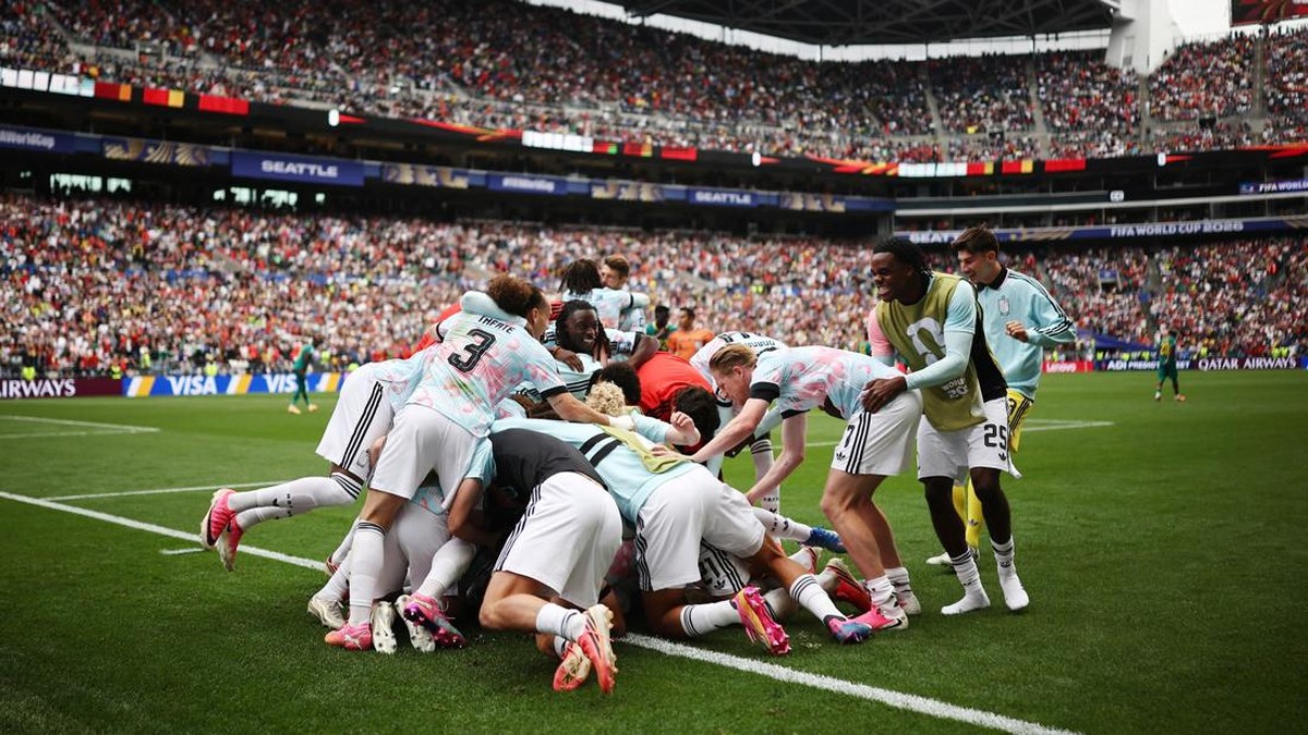 Soccer team in light-blue uniforms piles on each other celebrating a goal on the field as a full stadium cheers in the background and banners fly overhead.