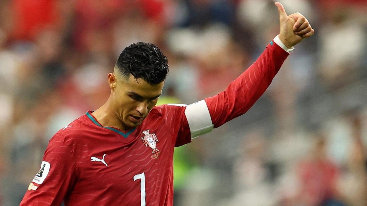 Portuguese soccer player in red jersey gives a thumbs-up on the field, captain's armband visible on the left arm.