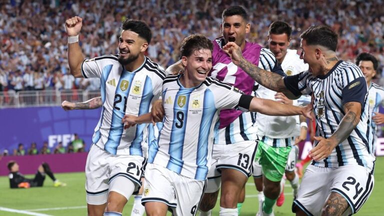 Argentina national team players celebrate a goal on the field, arms linked and smiling as fans cheer in the stadium behind them.