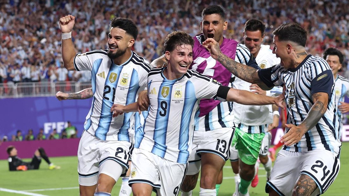 Argentina national team players celebrate a goal on the field, arms linked and smiling as fans cheer in the stadium behind them.