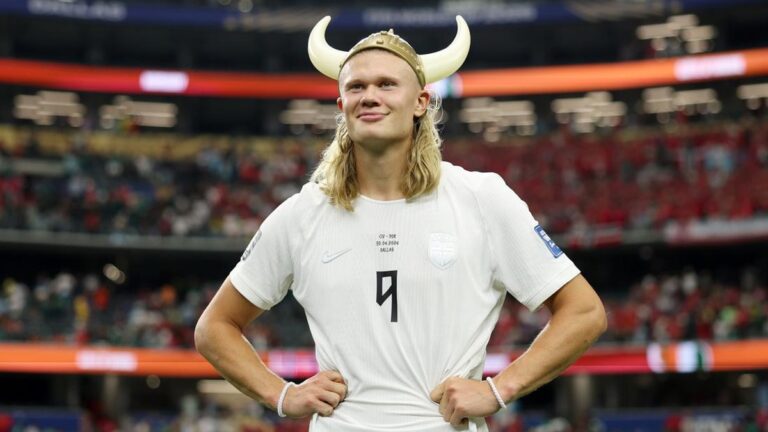 Soccer player in a white jersey with number 9 and a Viking horned helmet, hands on hips on a stadium field.