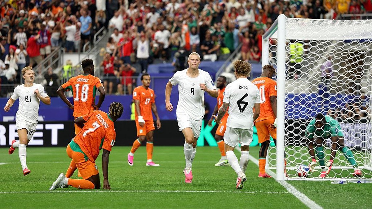 Soccer match near the goal: white-team players press forward while an orange-team defender kneels and the goalkeeper reaches for the ball by the net.