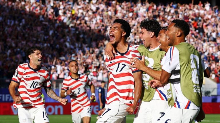 US men's soccer players in red-and-white striped jerseys celebrate a goal on the field as a crowded stadium cheers.