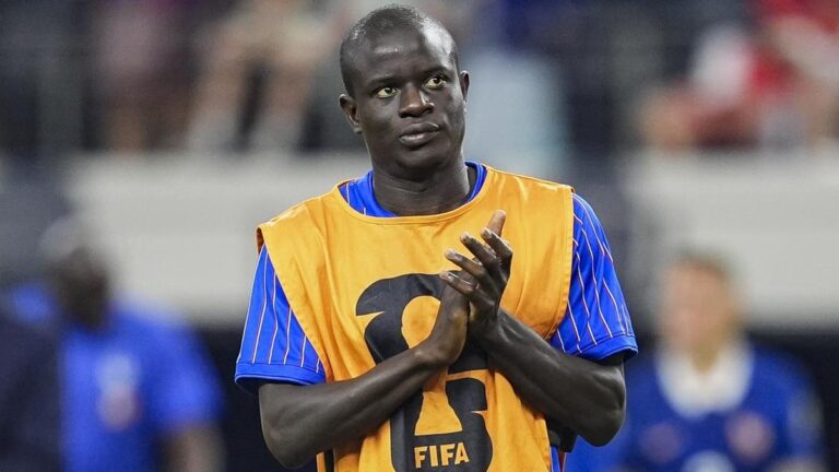 Football player wearing an orange bib with a FIFA logo stands on the field, hands held together in a prayer-like gesture.
