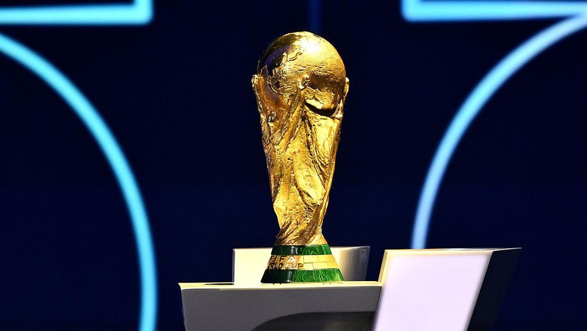 Golden FIFA World Cup trophy on a pedestal against a dark blue background, illuminated stage lighting visible behind it.