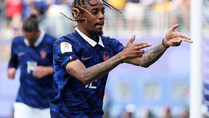 French footballer with braided hair in a blue France kit celebrates with arms outstretched on the pitch.