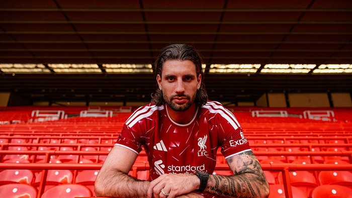 Athlete in a red Liverpool FC jersey sits with forearms on a red stadium seat, looking at the camera, tattoos on arms visible.
