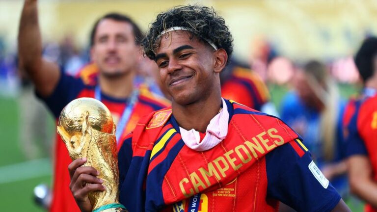 Soccer player smiling and holding a large gold trophy, wearing a red-and-blue champions sash that reads CAMPEONES in a celebratory stadium scene.