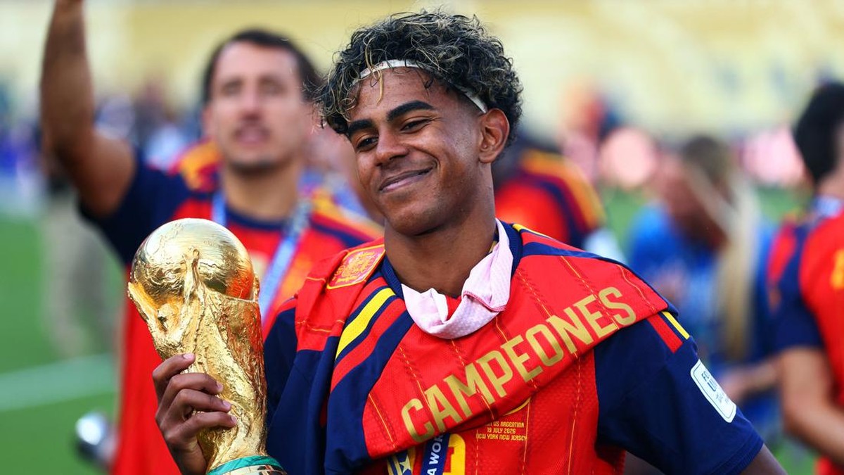 Soccer player smiling and holding a large gold trophy, wearing a red-and-blue champions sash that reads CAMPEONES in a celebratory stadium scene.