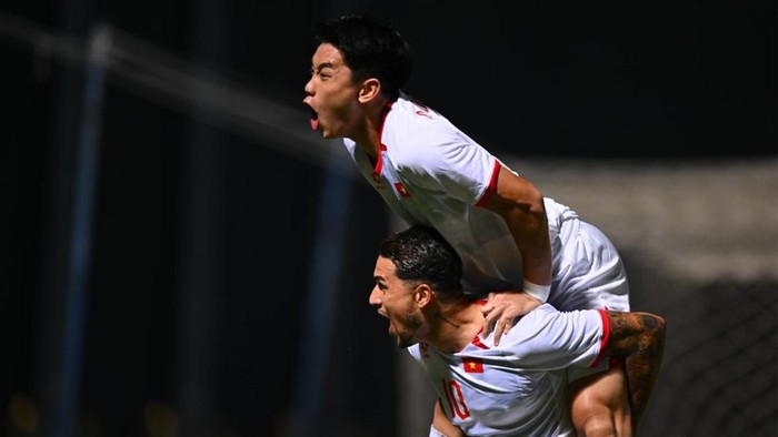 Two soccer players in white uniforms celebrate on the field, one carrying the other on his back in triumph.