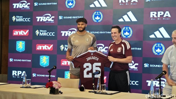 At a press conference, two men hold up a maroon football jersey that reads JAKARTA 25; a backdrop covered with sponsor logos is behind them.