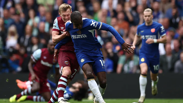 Two soccer players clash for the ball during a match; one in burgundy and light blue, the other in blue, in a crowded stadium.