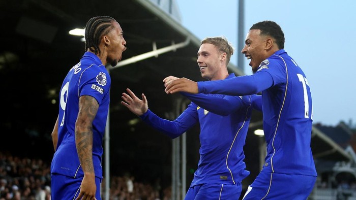 Three soccer players in blue uniforms celebrate together on the pitch near a stadium stands area.