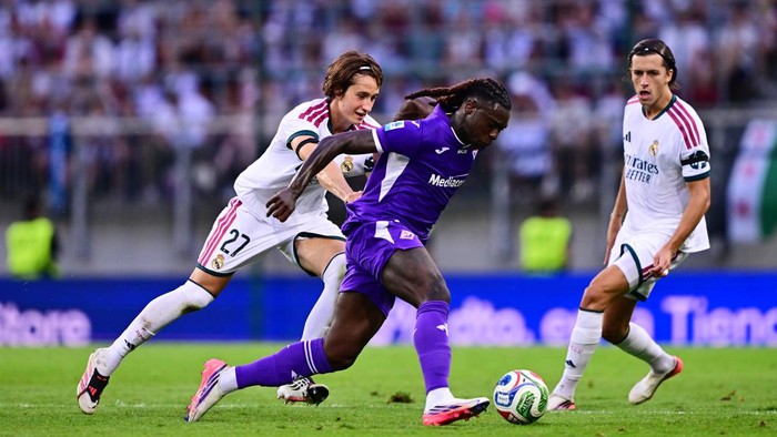 Soccer player in purple dribbles the ball past two opponents in white uniforms on a grass field.