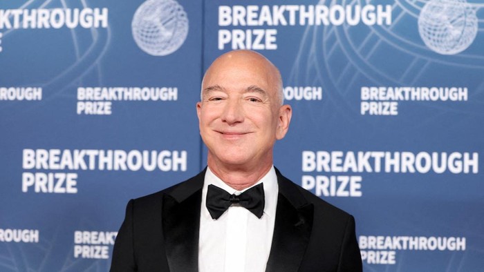 Smiling bald man in a black tuxedo with a bow tie stands in front of a blue backdrop that reads Breakthrough Prize.