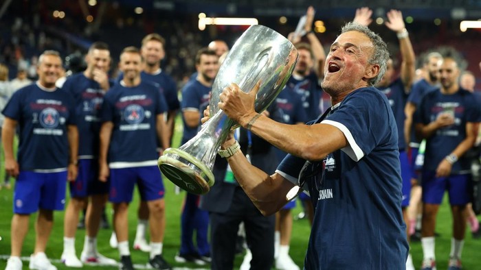Man in blue celebrates by kissing a tall silver trophy as teammates cheer in the background on a stadium field.