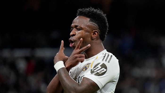 Real Madrid footballer on the field celebrating, wearing a white jersey, making a V sign with his fingers near his lips.