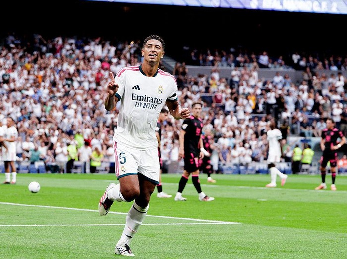 Cristiano Ronaldo in Real Madrid white kit celebrates a goal on a green soccer pitch with a full stadium behind him.