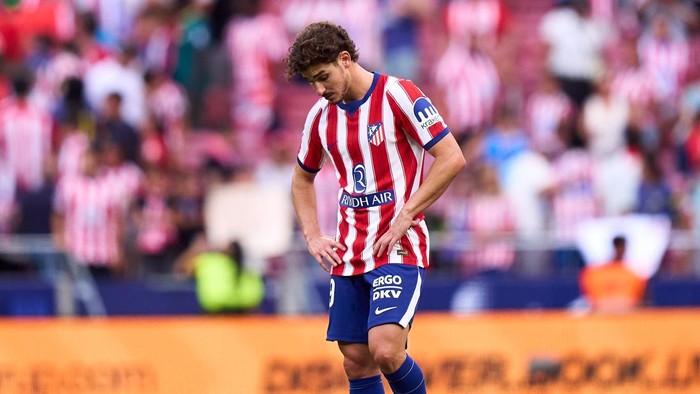 Soccer player in a red-and-white striped jersey with blue shorts standing on the pitch with hands on hips, crowd in the background