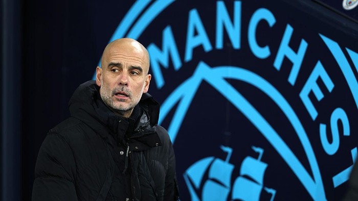 Man in a black jacket stands in front of a blue Manchester City logo backdrop, looking to the side.